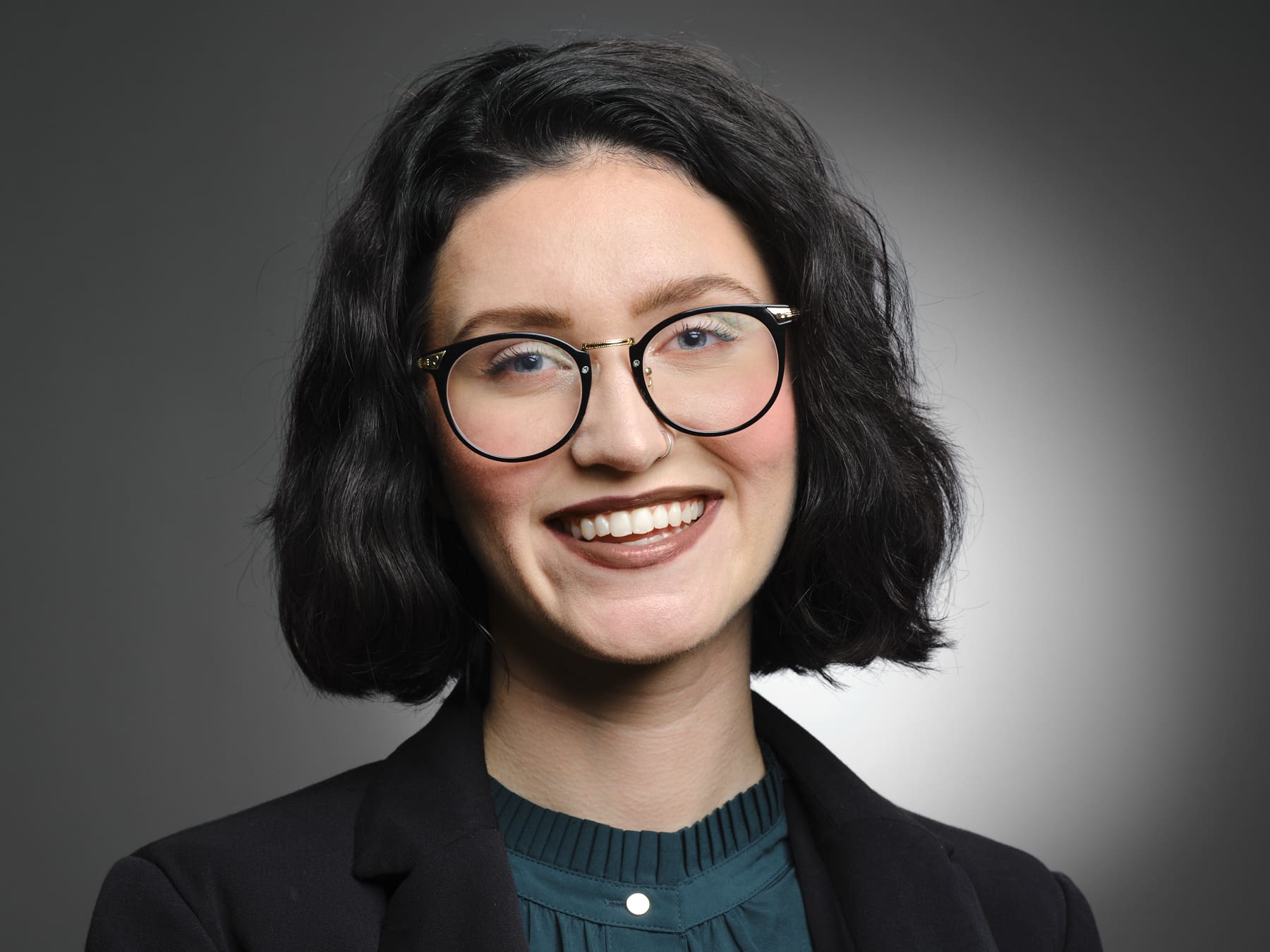 Professional woman with dark wavy hair and glasses smiling naturally in a black blazer against a gray studio backdrop.