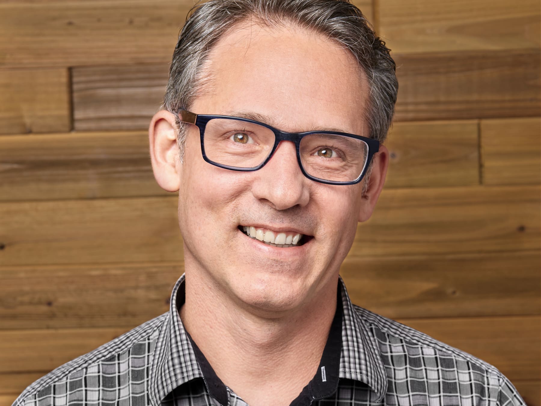 Professional headshot of a smiling man with gray hair wearing navy rectangular glasses and a gray checked shirt, photographed in front of a warm wood plank wall, with no glare on his lenses.