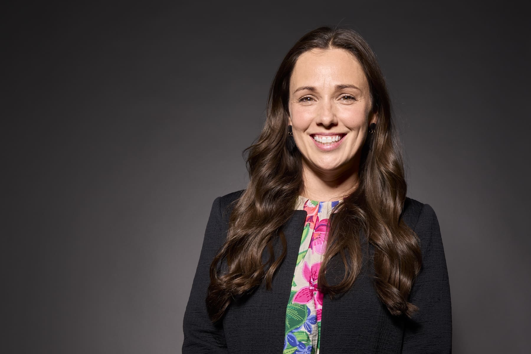 Confident professional headshot of a smiling businesswoman in a floral top against a dark studio backdrop