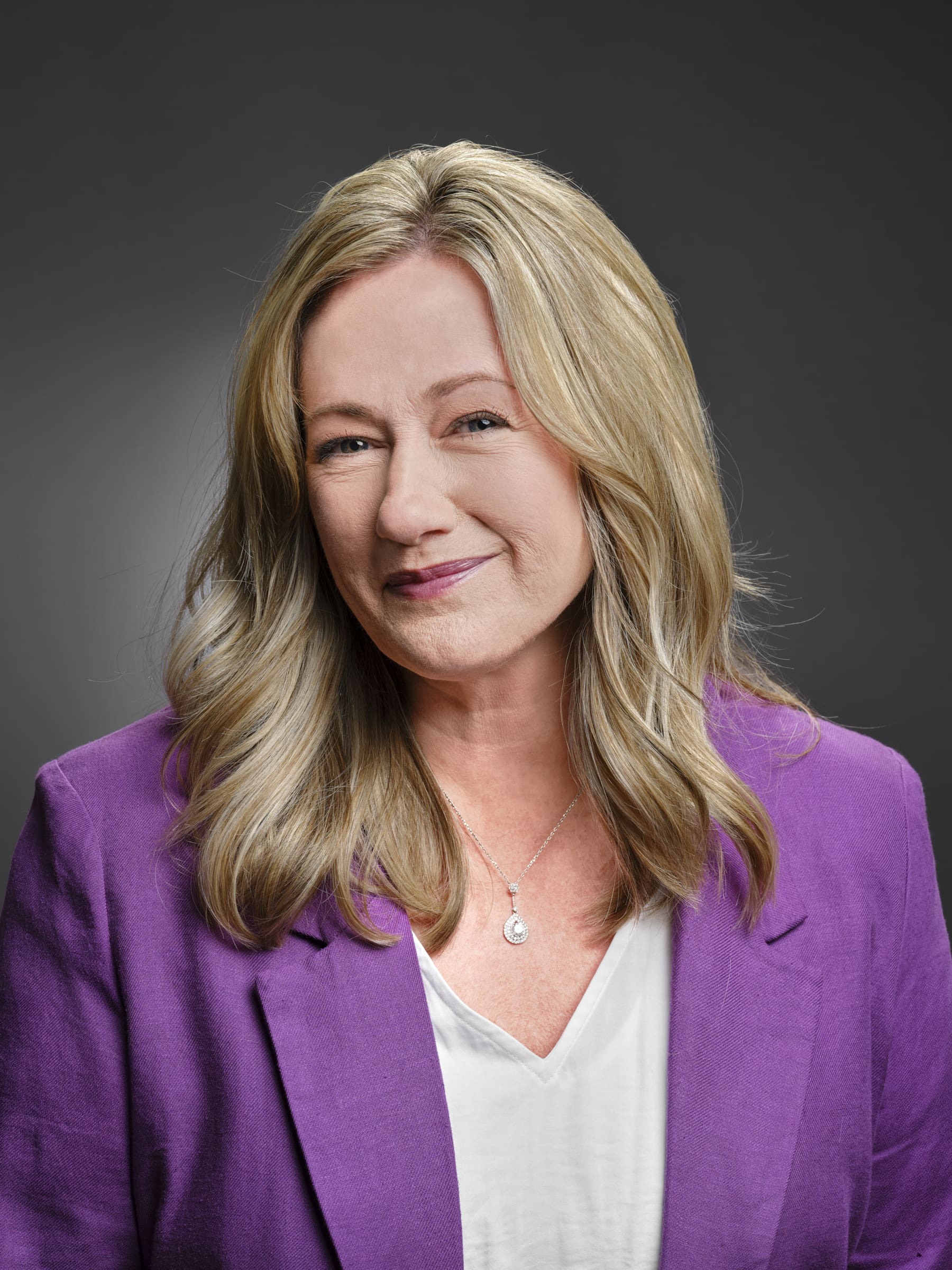 Professional headshot of a woman with long wavy blonde hair, wearing a purple blazer and white top, with a soft closed-mouth smile against a gray background
