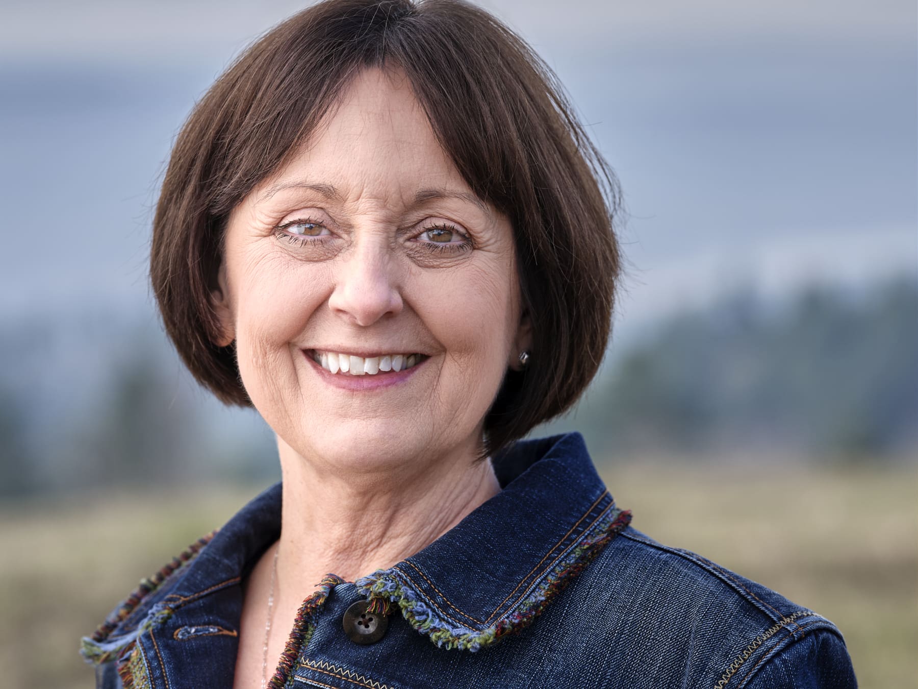 June Jones of Woodland Real Estate smiling at the camera in a dark denim jacket, with a soft outdoor background of trees and hills.